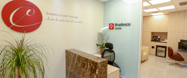 Sala de recepção com balcão em mármore, com aromatizador de ambiente e cadeira de escritório atrás do balcão. Ao fundo, há a sala de atendimento com frigobar, cafeteira e uma poltrona de couro, na cor bege. Na sala, também há uma mesa de vidro com computador e cadeira de escritório.