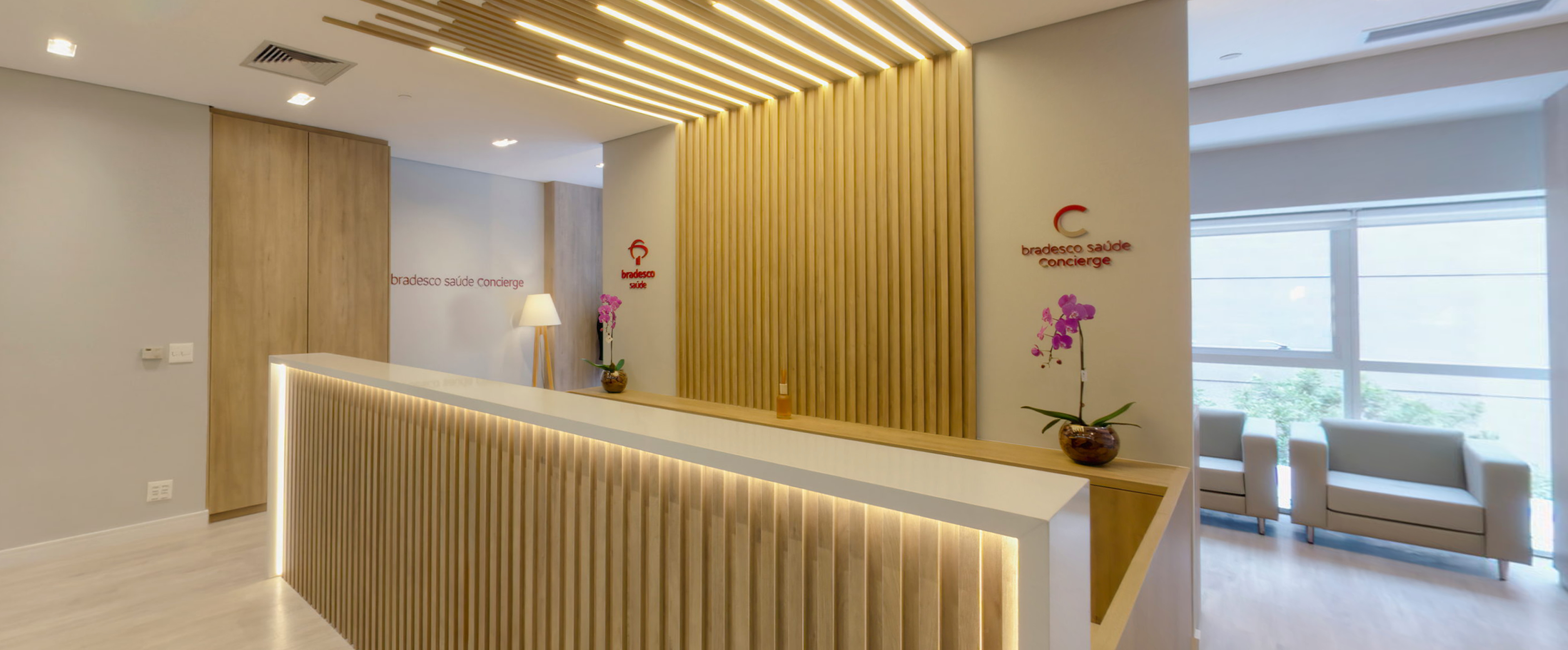 Sala com 4 poltronas decorativas de couro, na cor branca. No meio da sala, há uma mesa de centro, com revistas e vaso decorativo. Também há uma televisão em frente às poltronas, e plantas harmonizando com o ambiente. Ao fundo, uma mesa de recepção, com computador e cadeira de escritório.